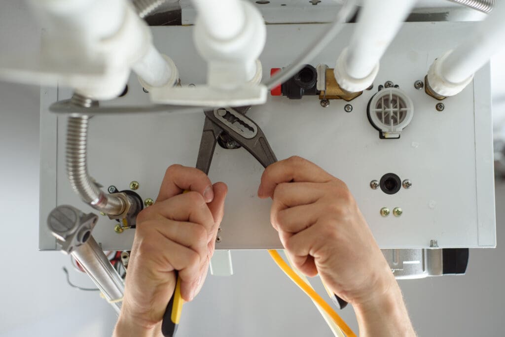 A skilled gas engineer uses pliers to adjust pipes beneath a boiler. Various pipes, connectors, and valves suggest ongoing maintenance or repair work. The background is blurred, highlighting the hands and tools involved in ensuring efficient gas central heating.
