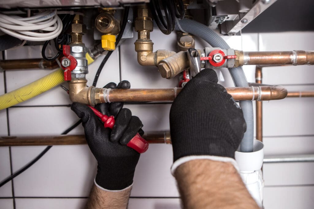 A person wearing black gloves expertly wields a red pipe wrench to adjust copper plumbing pipes connected with brass fittings and valves, all crucial for heating system repairs. Other pipes are visible against the backdrop of a white tiled wall.