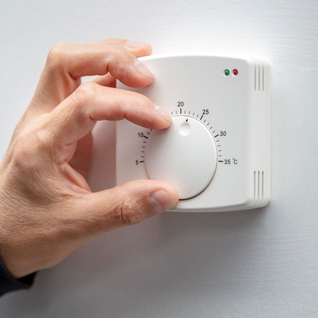 A person adjusts a white analog thermostat mounted on a wall. The hand is turning the round dial clockwise, changing the temperature setting. The thermostat displays temperatures from 5 to 35 degrees Celsius.