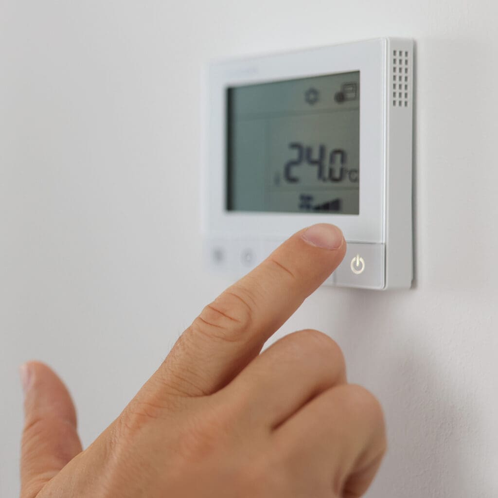 Close-up of a hand adjusting a wall-mounted digital thermostat displaying 24°C. The wall is white, and the room has a minimalistic design with soft lighting.