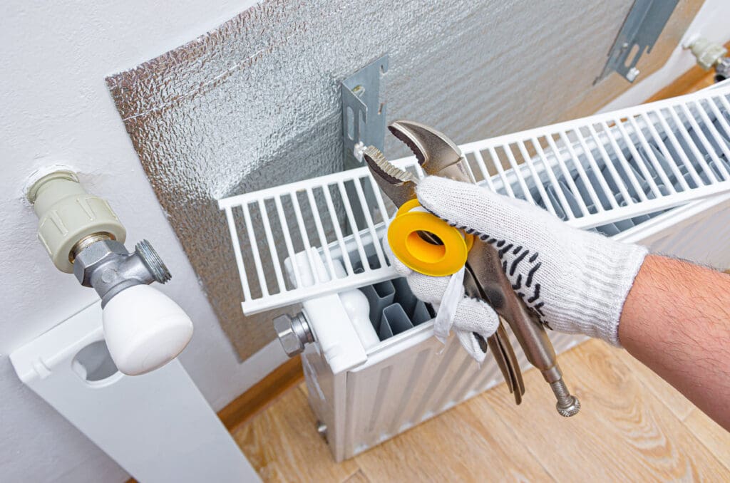 A person wearing a glove holds a wrench and a roll of tape above a radiator. The radiator is positioned against a wall, with a shiny insulating panel behind it. A valve is visible on the left side of the radiator.
