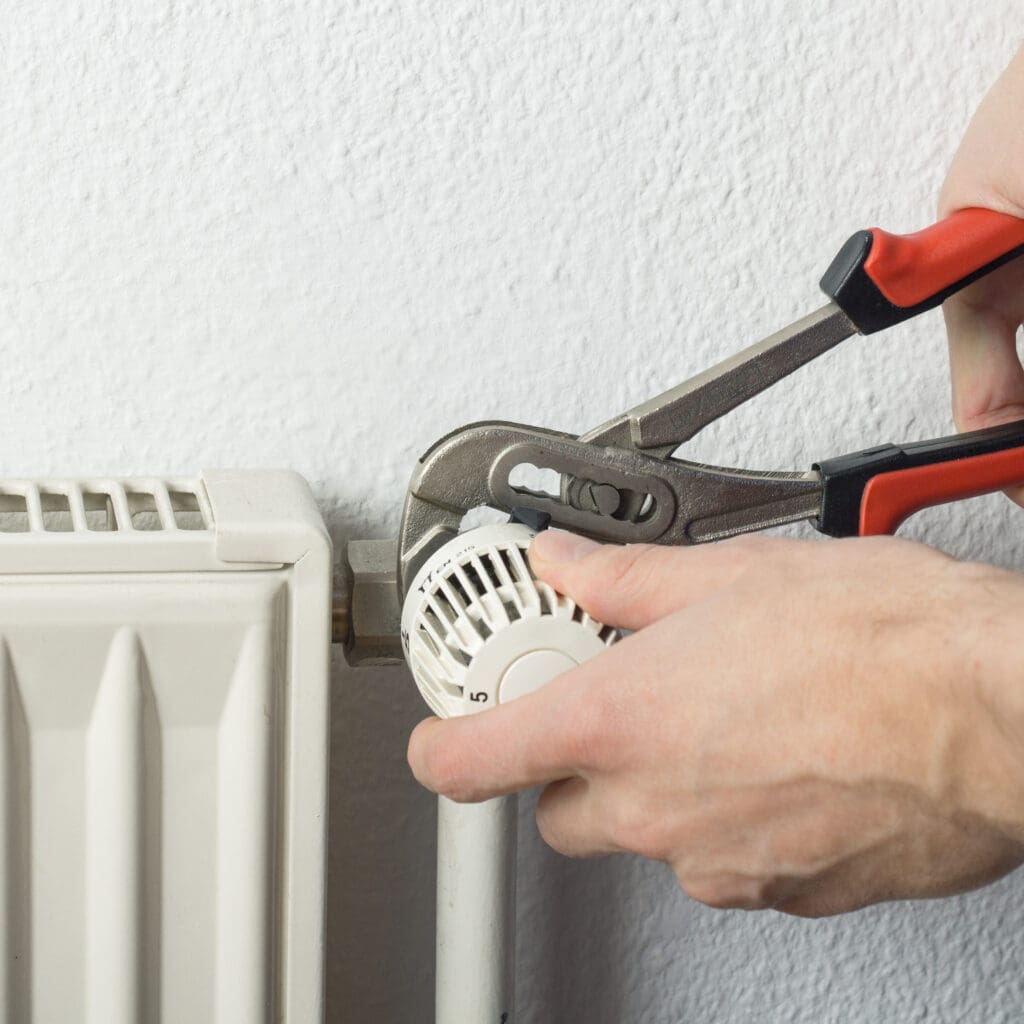 A person’s hands use pliers to adjust or repair the thermostat valve on a white radiator attached to a wall.