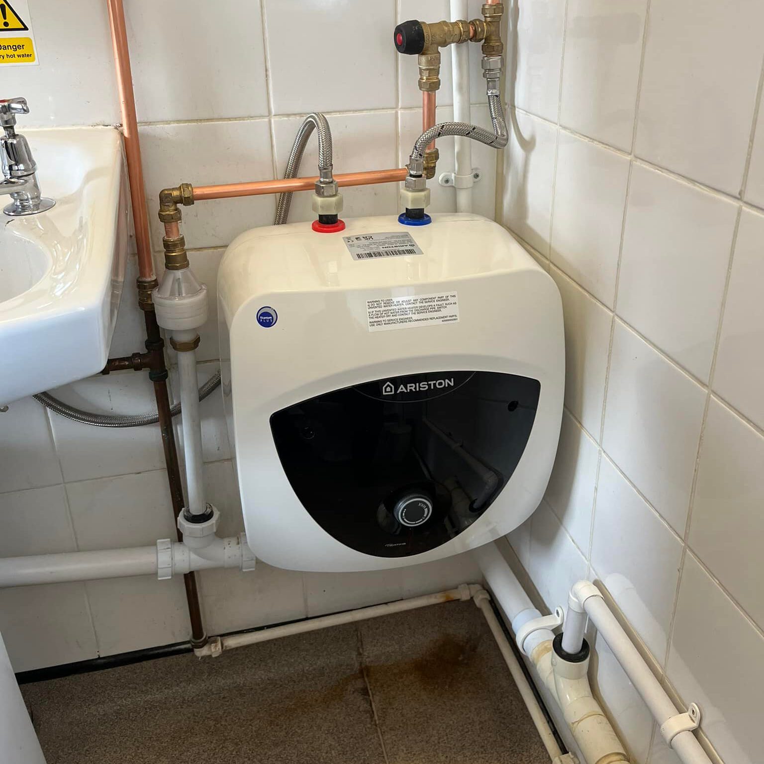 A small white Ariston water heater, part of a recent boiler upgrade, is installed in a tiled corner with visible pipes and valves. Positioned next to a white sink on the gray floor, it sits beneath a safety warning sign on the wall.