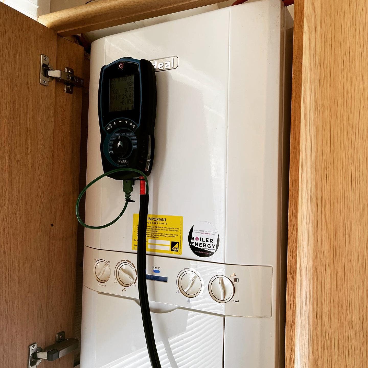 A white boiler unit, recently upgraded, is installed inside a wooden cabinet. The boiler, part of a modern gas central heating system, has a digital device attached by a cable. Control knobs and labels adorn the front as sunlight filters through partially opened blinds nearby.
