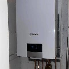 A white Vaillant boiler mounted on a wall, displaying a digital screen with a temperature of 34°C. Copper pipes are connected at the bottom. The boiler is installed in a utility or basement room.