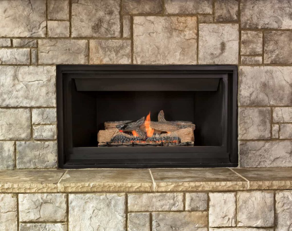 A gas fireplace with artificial logs burning inside a black frame, set in a stone wall with large, rectangular stone tiles.