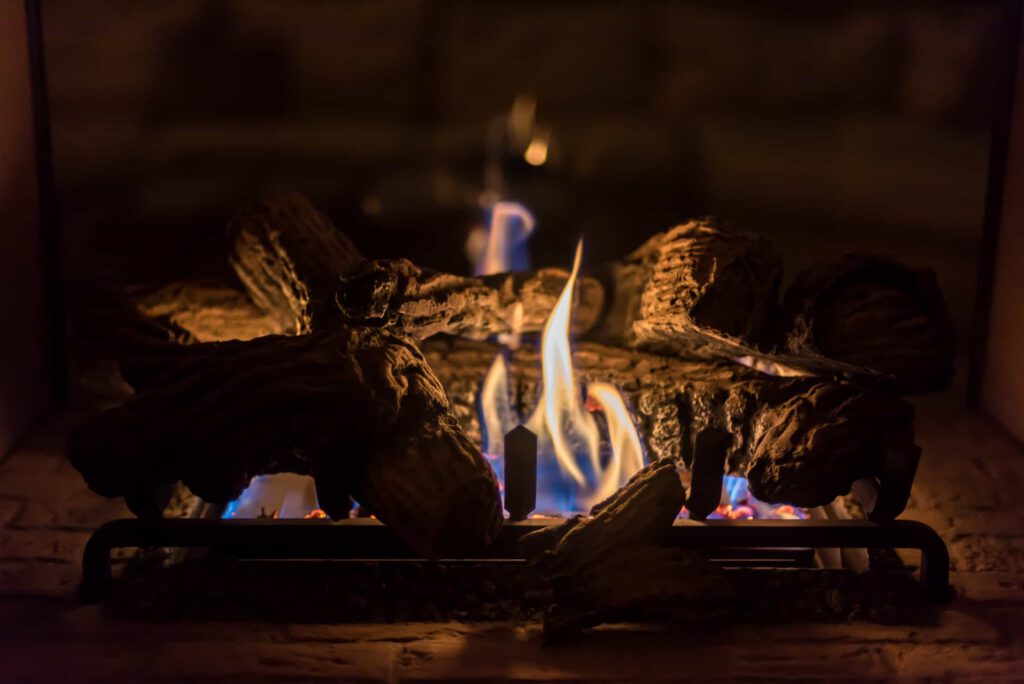 A close-up of a lit fireplace with logs burning, emitting yellow and blue flames. The firewood rests on a metal grate, creating a warm and cozy atmosphere.