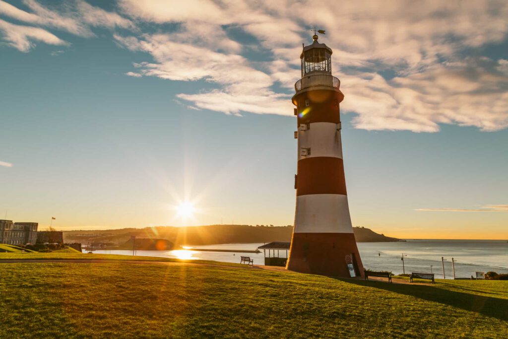 A tall red and white striped lighthouse stands on a grassy hill by the sea at sunrise, with sunlight casting long shadows and clouds scattered across the sky.
