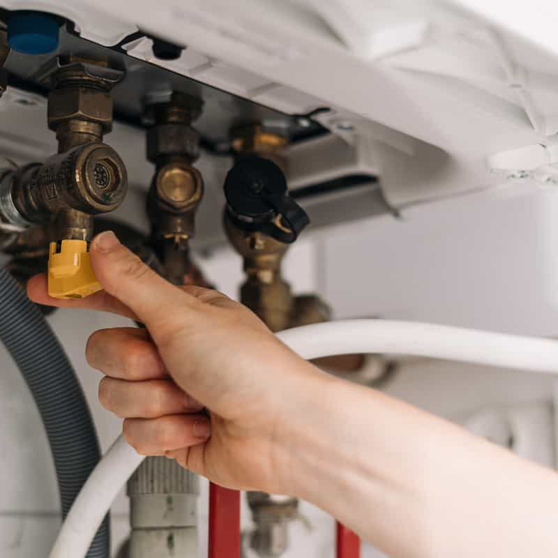 A close-up of a hand turning a yellow valve under a sink or appliance, with pipes, hoses, and metal fittings visible in the background.