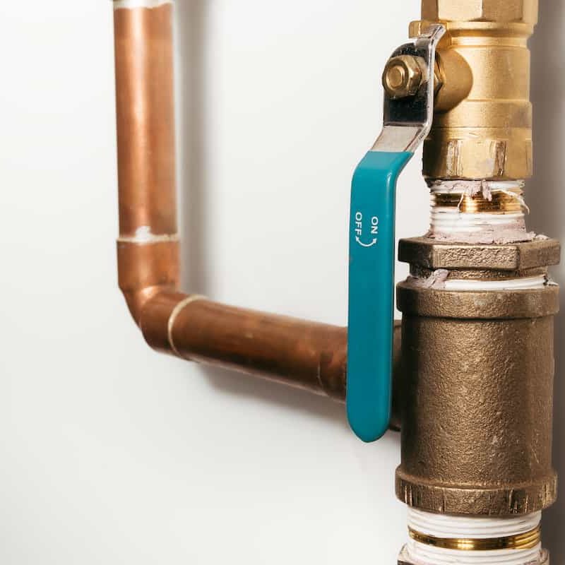 Close-up of a metal pipe with a brass valve and a turquoise handle showing "ON" and "OFF" positions. The pipe is attached to a white wall.