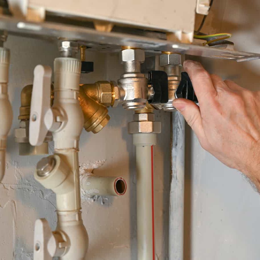 A person’s hand is adjusting a valve on a network of metal and plastic pipes attached to a wall, likely part of a home heating or plumbing system.