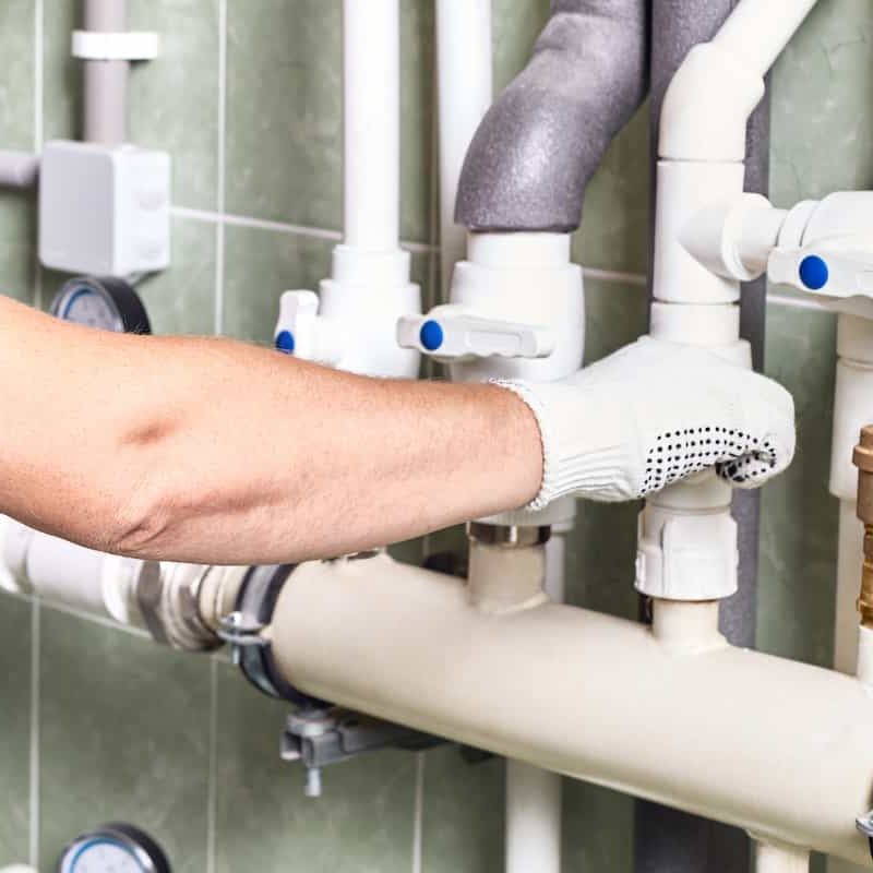 A person wearing a white work glove adjusts a valve on a network of white pipes and fittings, mounted on a green tiled wall, possibly as part of a plumbing or heating system.