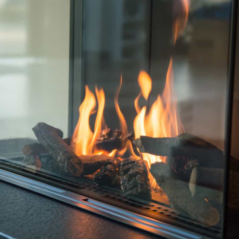 A close-up of a modern glass-enclosed fireplace with burning logs and visible yellow-orange flames.