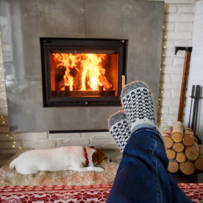 A person wearing patterned socks relaxes with feet up near a fireplace, while a dog sleeps on a rug in front of the fire. Stacked firewood and fireplace tools are nearby.
