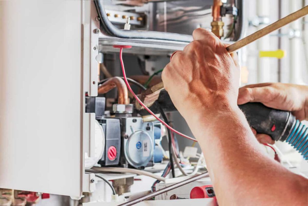 A person’s hands are repairing or performing maintenance on the internal components of a gas boiler, using tools to adjust wires and pipes inside the open unit.