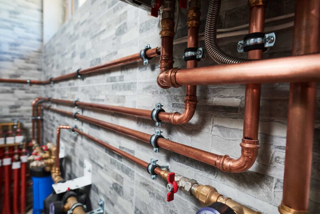 Copper pipes neatly arranged along a tiled wall, secured with metal brackets, featuring several valves and connectors as part of a building’s plumbing or heating system.