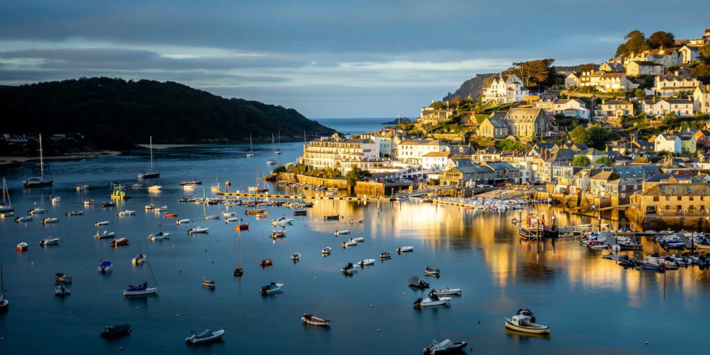 A picturesque harbor at sunset with numerous boats on calm water, and a hillside town with white and beige buildings bathed in golden sunlight, surrounded by lush greenery and a distant view of the sea.