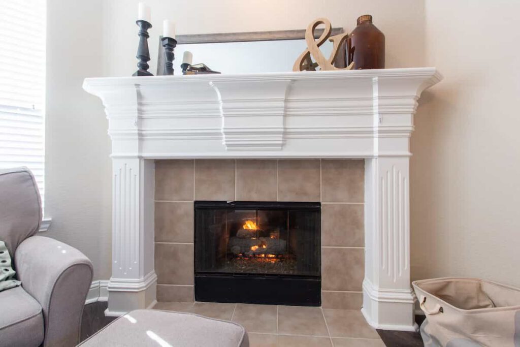 A lit fireplace with a white mantel decorated with candles, a mirror, and vases. An armchair, an ottoman, and a basket are nearby, creating a cozy living room setting.