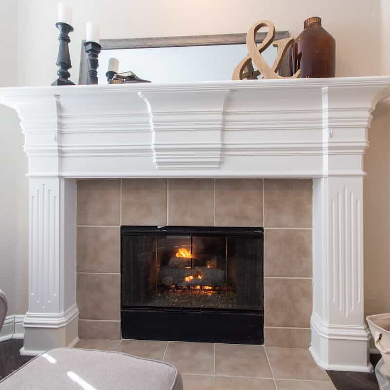 A white fireplace mantel decorated with candles, a mirror, and pottery sits above a tile-surrounded fireplace with a burning fire inside.
