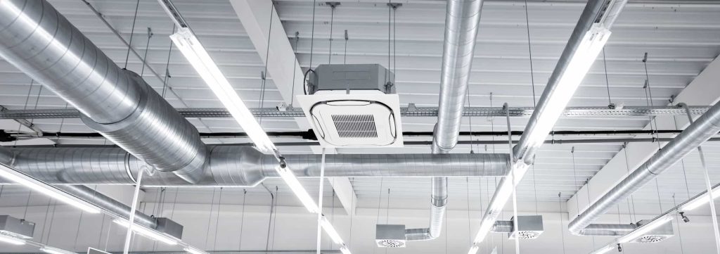 Ceiling view of a modern industrial building showing metal air ducts, a ceiling-mounted HVAC unit, and rows of bright fluorescent light fixtures.