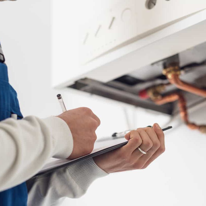 A person holding a clipboard and pen inspects a gas boiler, checking components and making notes. Copper pipes and part of the boiler unit are visible in the background.