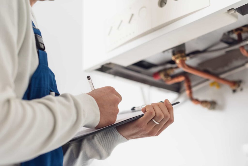 A person in blue overalls holds a clipboard and pen, inspecting and taking notes on exposed pipes and components of a gas boiler or heating system.