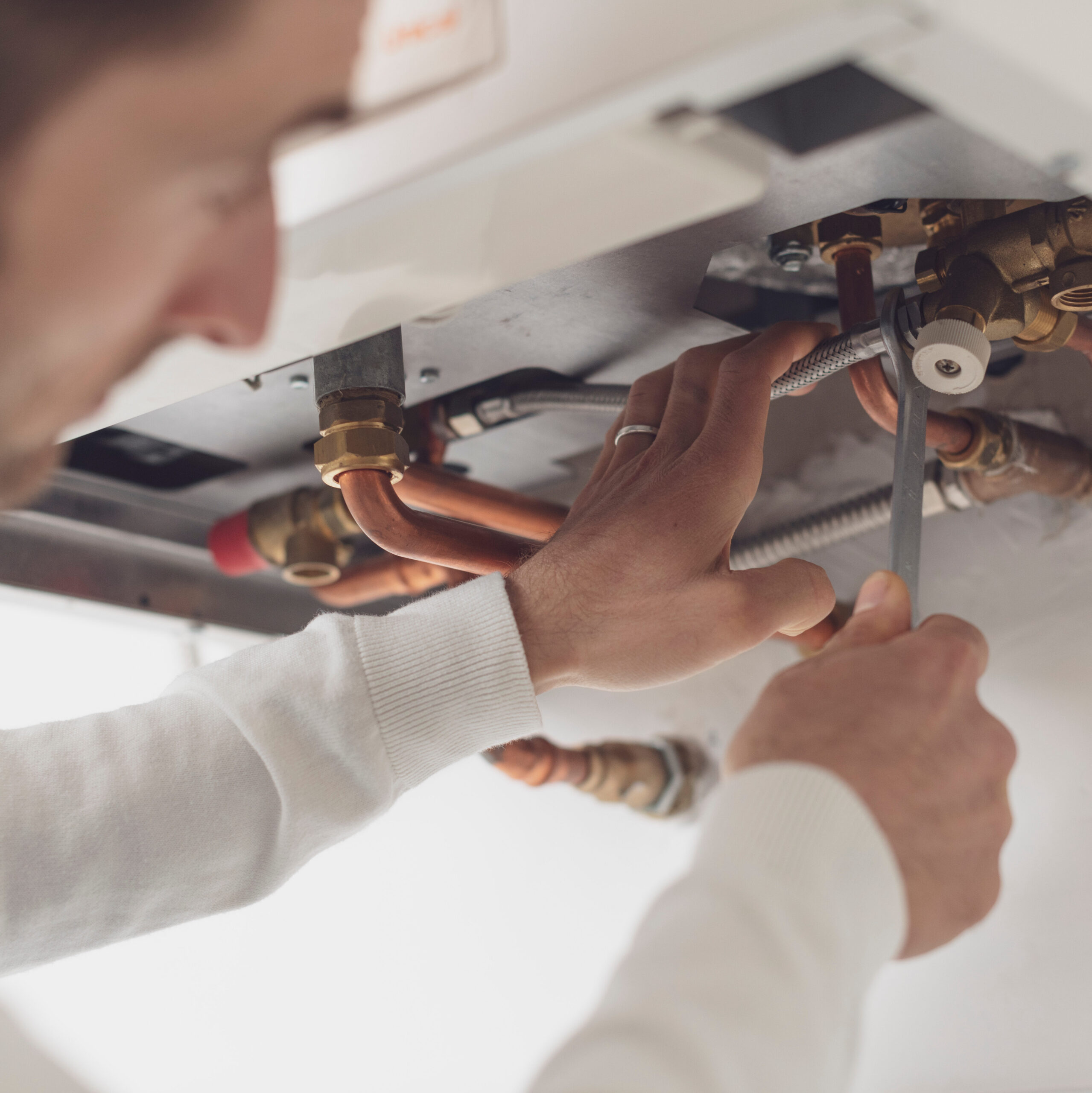 A person in a white shirt is using a wrench to work on copper pipes beneath a boiler or appliance. The focus is on the hands and tools, with the person's face slightly blurred in the foreground.
