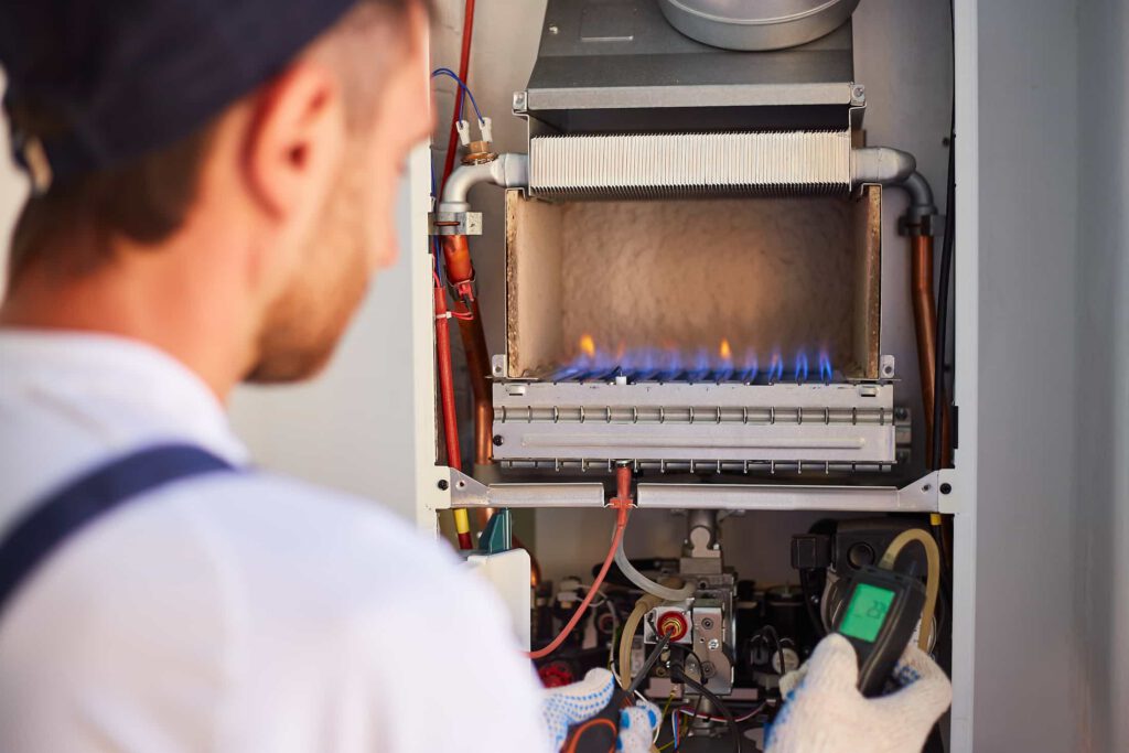 A technician wearing gloves inspects and tests a gas boiler using diagnostic equipment, while blue flames are visible inside the boiler. The technician’s face is blurred in the foreground.