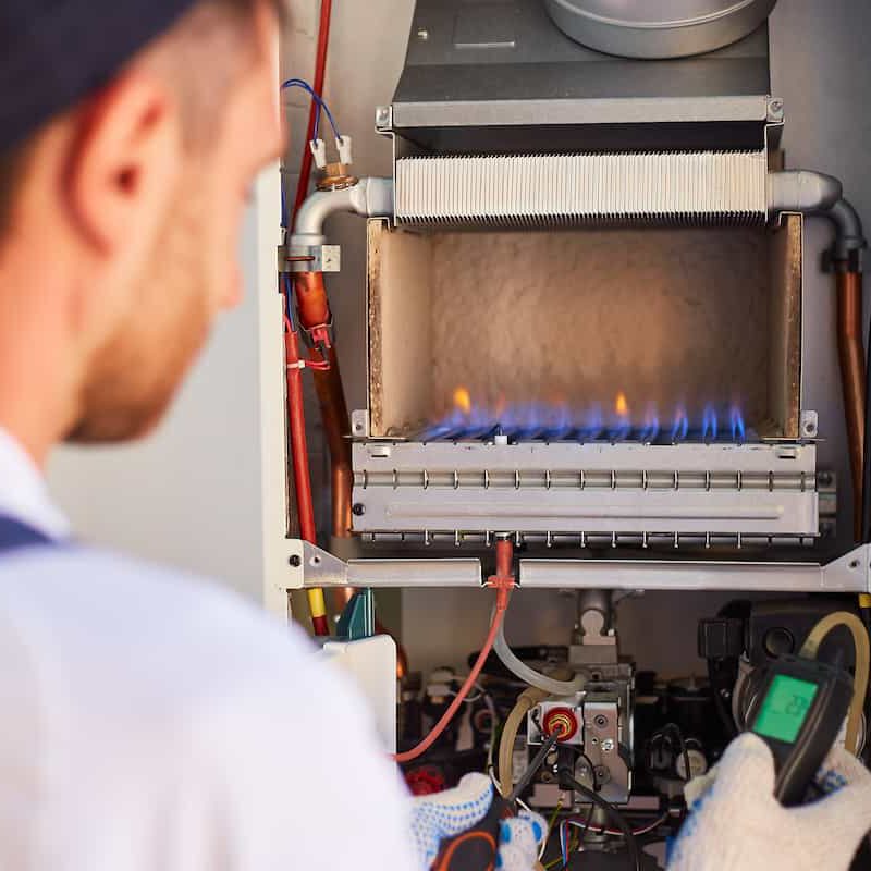 A technician wearing gloves inspects and tests a gas boiler with blue flames inside, using a digital device. The boiler's inner components and pipes are visible.