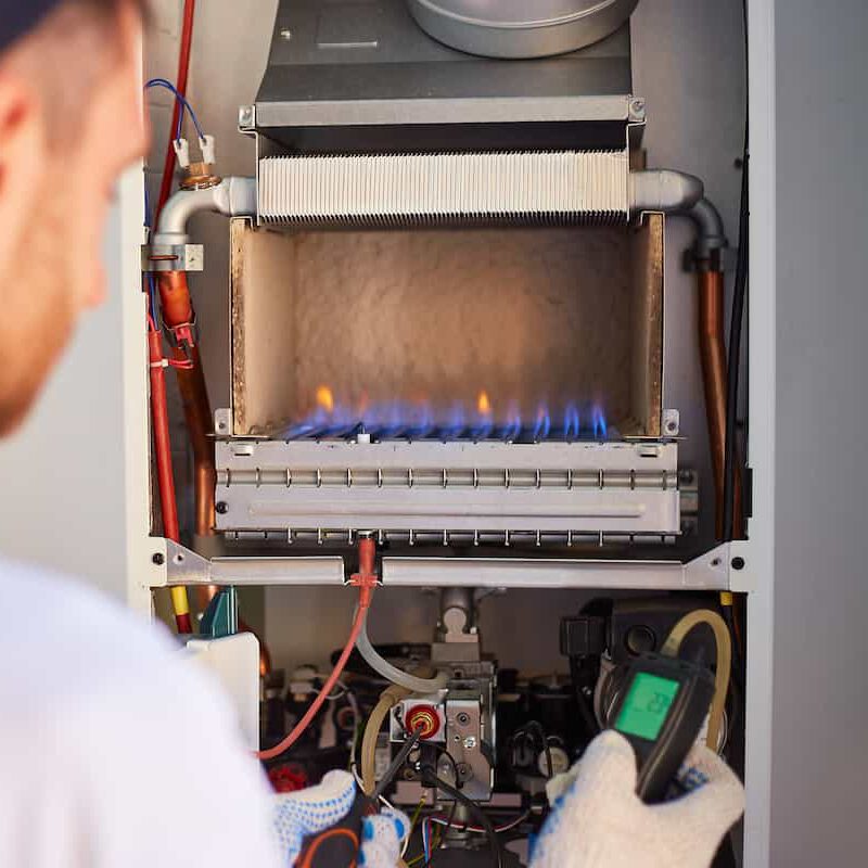 A technician in gloves uses a diagnostic tool to inspect a gas boiler with visible blue flames inside, checking its operation and safety.