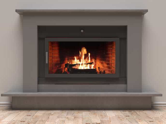 A modern, gray-framed fireplace with a glass front shows a burning fire inside, with visible flames and logs. The fireplace is set against a light-colored wall above a wooden floor.