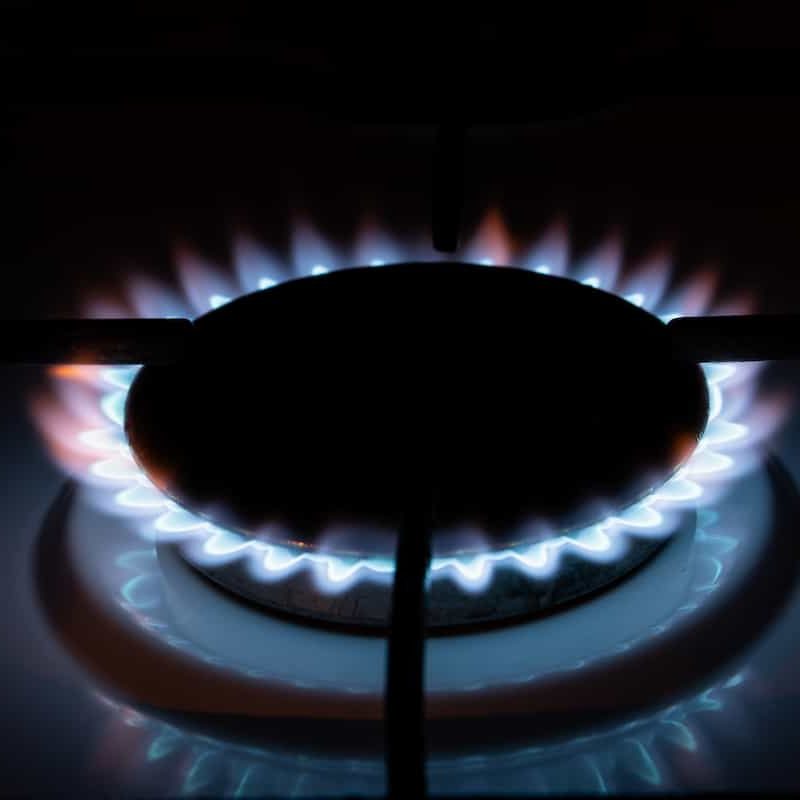 A close-up of a lit gas stove burner with blue flames evenly surrounding the dark burner grate in a dimly lit setting.