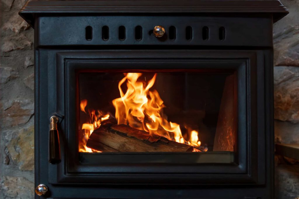 A close-up of a black wood-burning stove with visible orange flames and burning logs inside, set against a stone wall background.