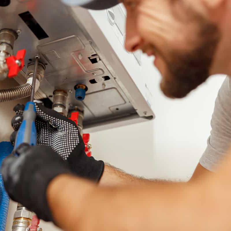 A smiling plumber wearing gloves uses a screwdriver to repair a metal appliance, possibly a water heater or boiler, with visible pipes and valves.