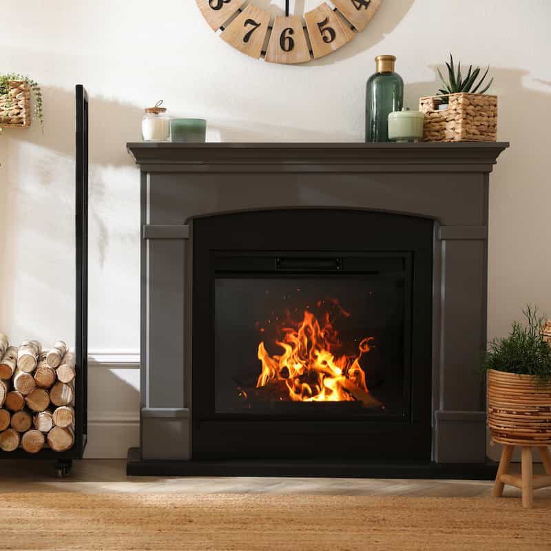 A modern gray fireplace with a fire burning inside, surrounded by decorative plants, stacked firewood, and green glass vases. A large wall clock hangs above the mantel.