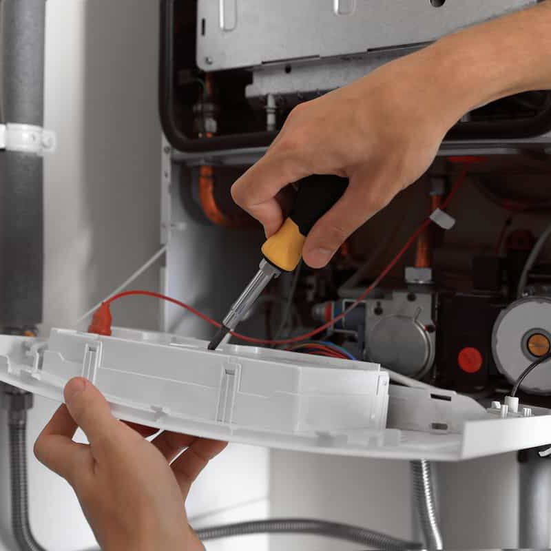 A person uses a screwdriver to repair or adjust the internal components of a furnace or boiler, with various wires and mechanical parts visible.