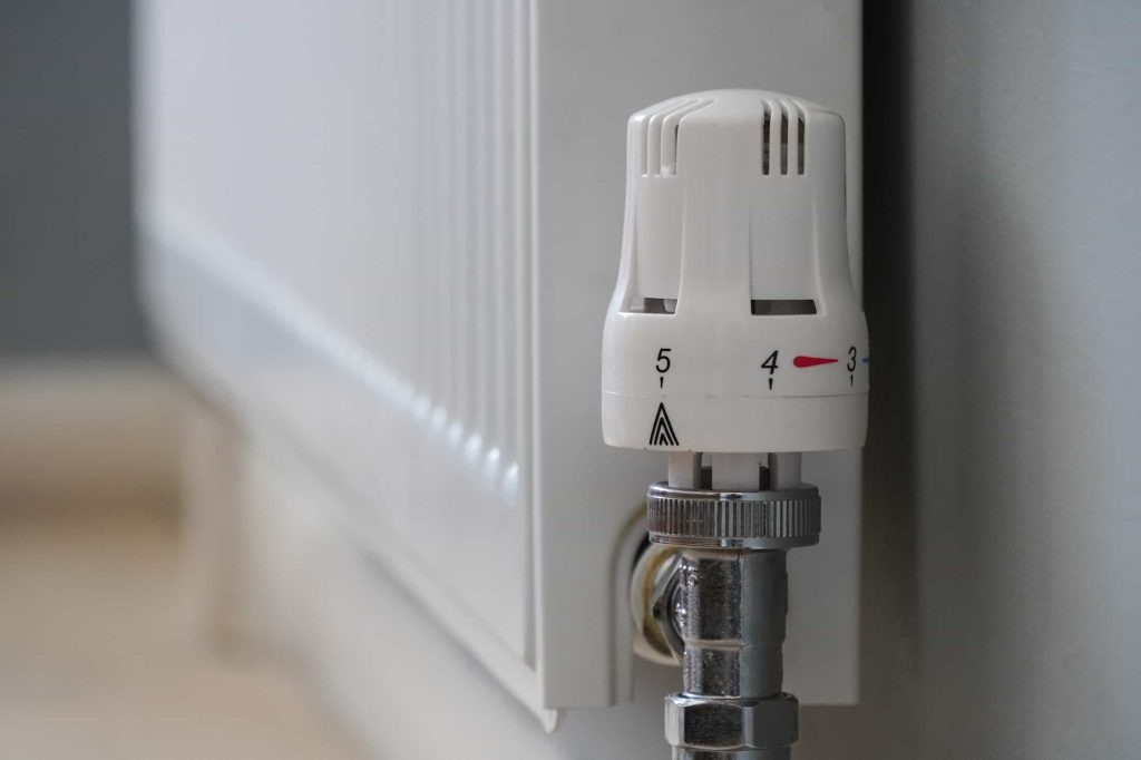 Close-up of a white radiator with a temperature control valve set between 3 and 4, attached to a wall, with part of the pipe and floor visible.