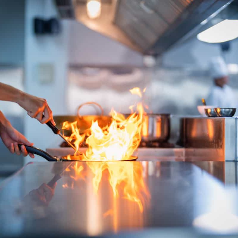 A chef’s hand holds a pan over a stove with flames rising from it in a professional kitchen, with pots and a blurred figure in the background.