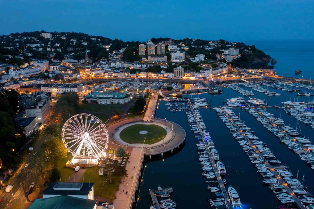 Aerial view of a marina at dusk with many docked boats, a large illuminated Ferris wheel, pathways, and surrounding town buildings with lights reflecting on the water.