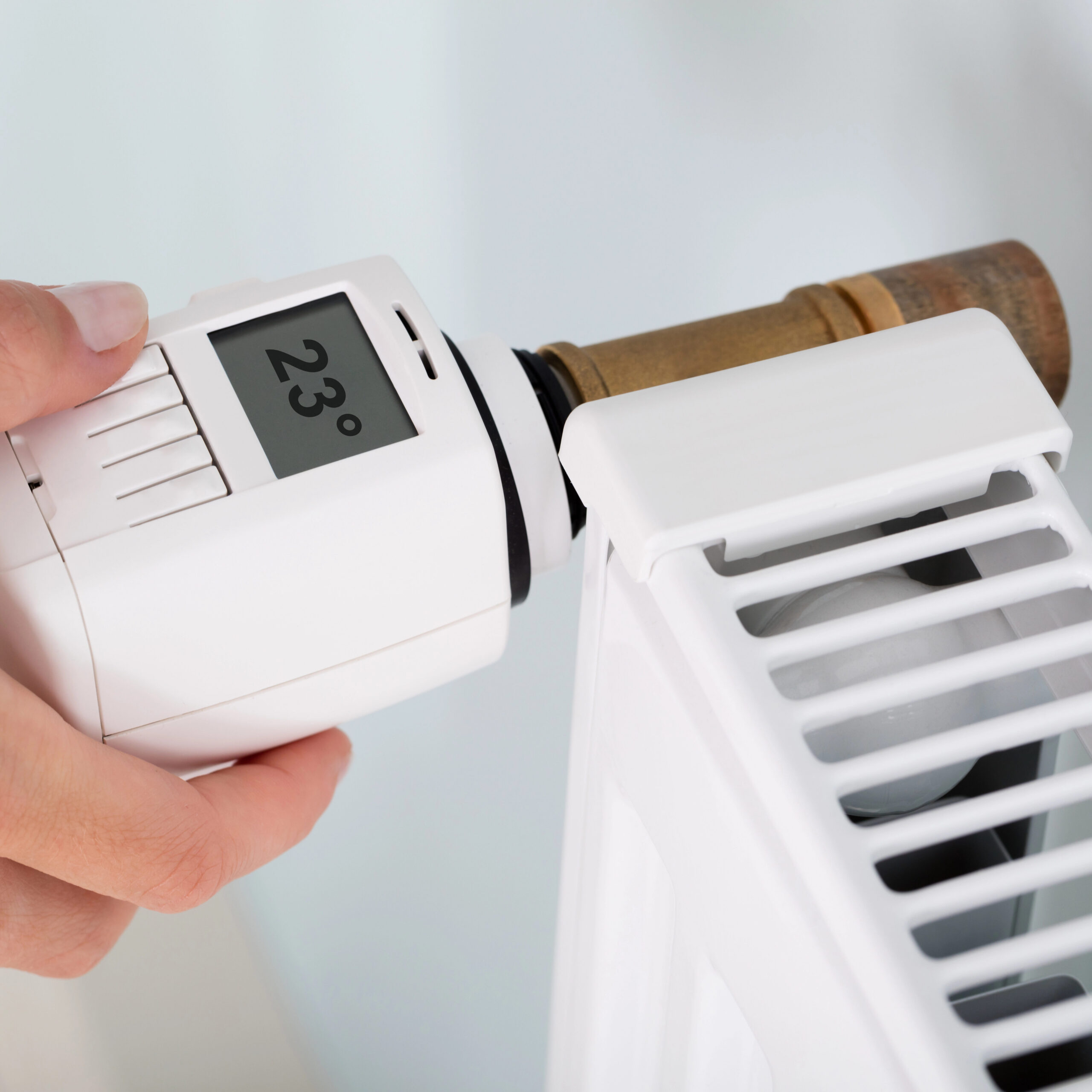 A hand adjusts a digital thermostat attached to a radiator, set at 23 degrees, indicating precise temperature control in the gas central heating system. The white radiator stands out against the softly blurred background.